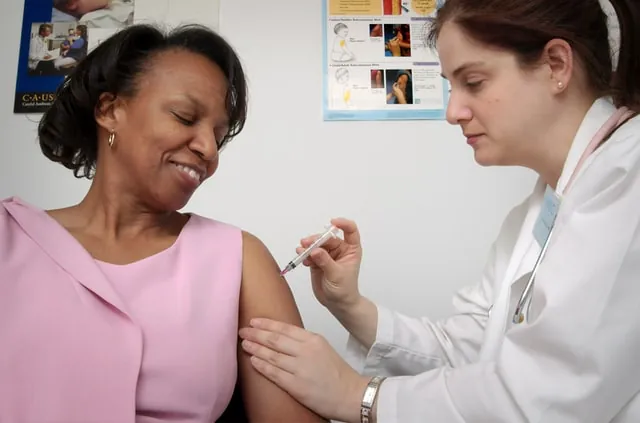 A health care worker in a white coat gives an injection in the upper arm of a smiling woman in a pink top.