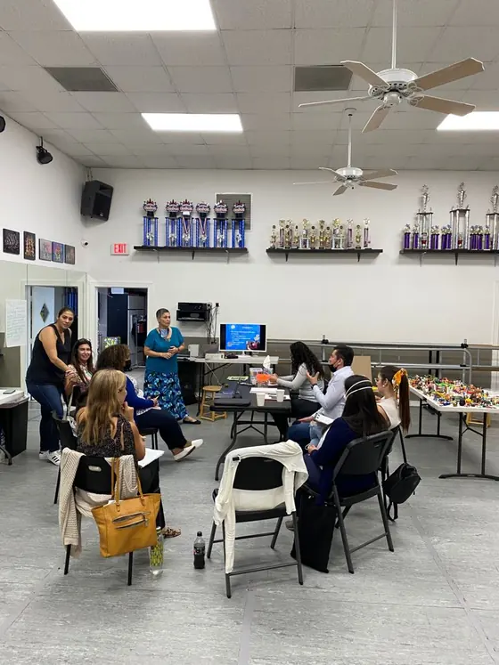 Therapists sit on folding chairs in a large room during the play therapy training while a woman in a teal top stands and speaks; a screen shows a slide and a long table holds small toy figures.