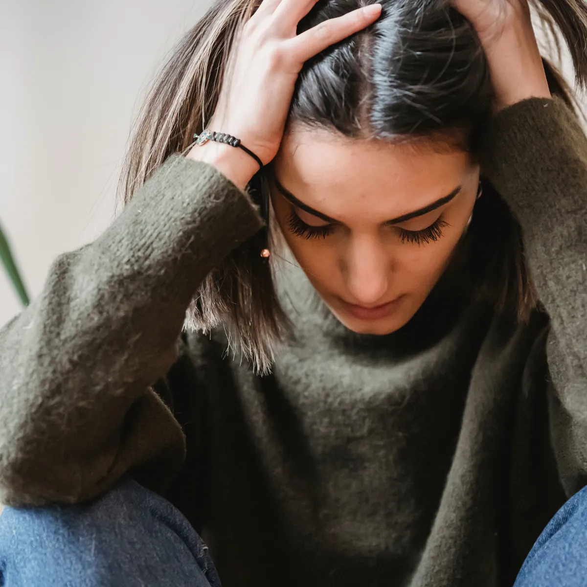 A young woman with long dark hair and a green sweater sits with her head down and both hands pressed to her head, looking distressed.