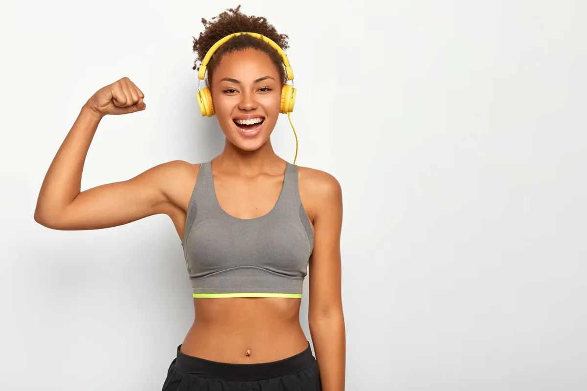 A smiling woman with curly hair, wearing yellow headphones and a gray sports top, flexes her arm against a white background.