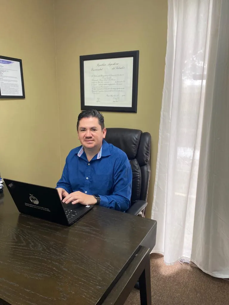 A smiling man in a blue shirt sits at a laptop at a dark wooden desk in an office, with a framed diploma on the wall behind him.