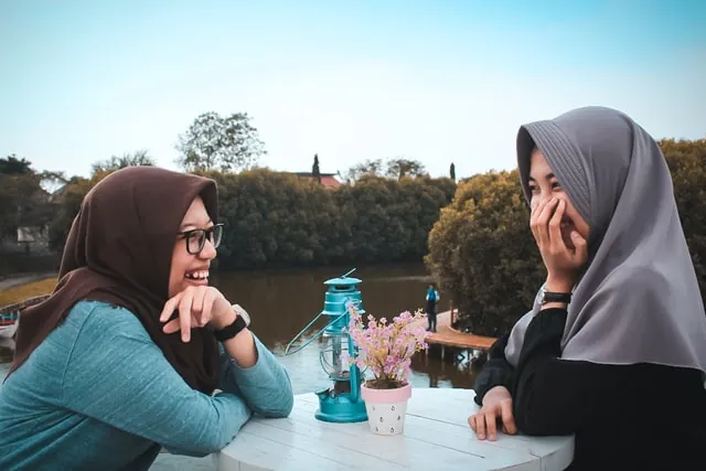 Two young women in headscarves laugh together across a small table by a lake, with a potted flower and a lantern between them.