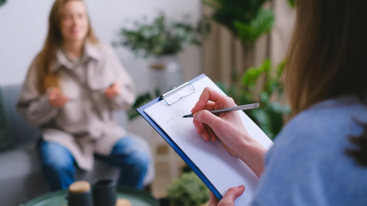 Over the shoulder of a therapist taking notes on a clipboard while a smiling woman on a sofa talks in a bright room with plants.