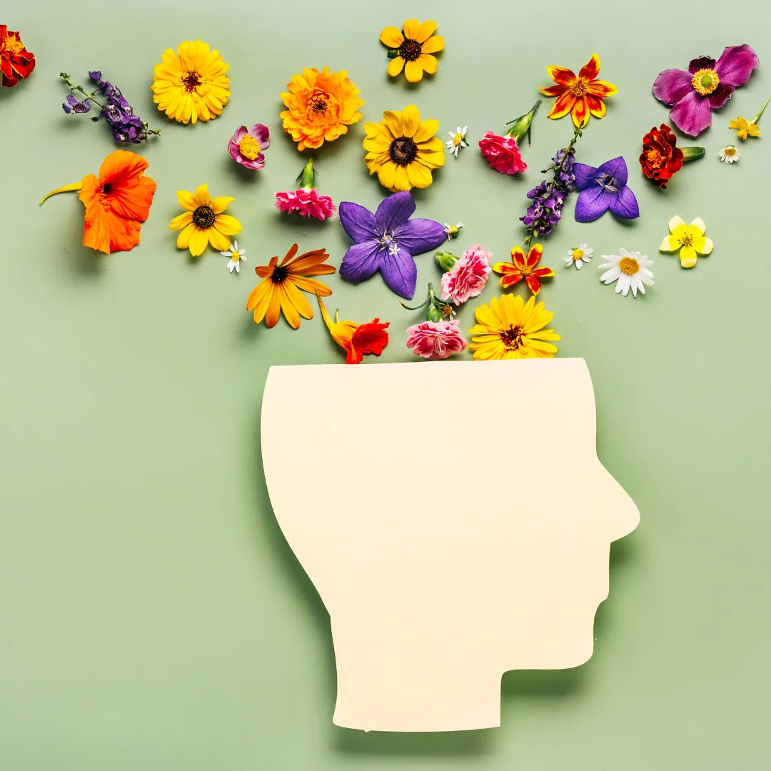 A cream paper cutout of a head in profile on a sage green background, with colorful flowers spilling out of the top of the head.