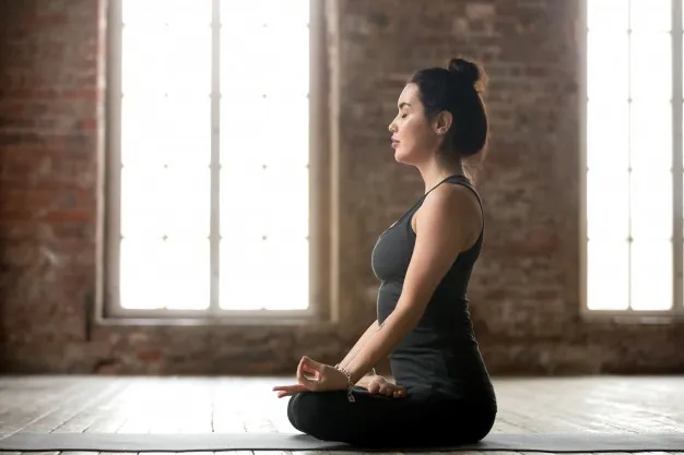Young woman meditating in a cross-legged yoga pose with her eyes closed, in a bright studio with brick walls and tall windows